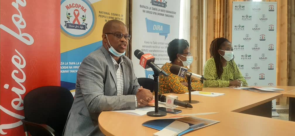 NACOPHA leaders at a press conference, seated beneath the NACOPHA banner and the organisation's vision statement, speaking into television and radio microphones